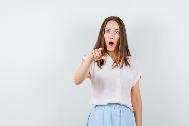 Jeune fille pointant la caméra en t-shirt, jupe et regardant étonné, vue de face.