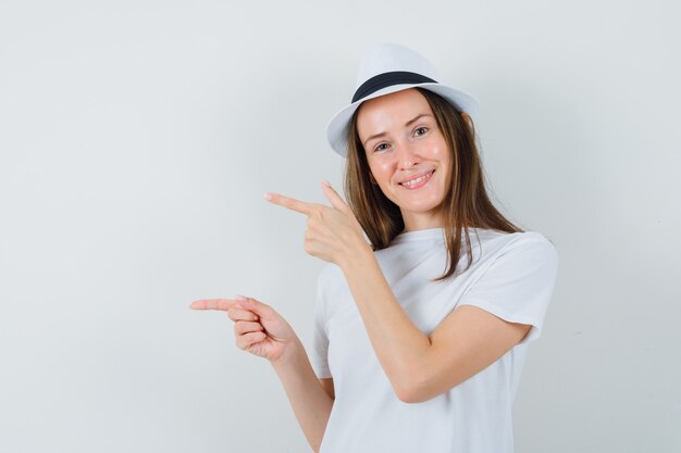 Jeune fille pointant les doigts vers le côté gauche en t-shirt blanc, chapeau et à la jolly. vue de face.