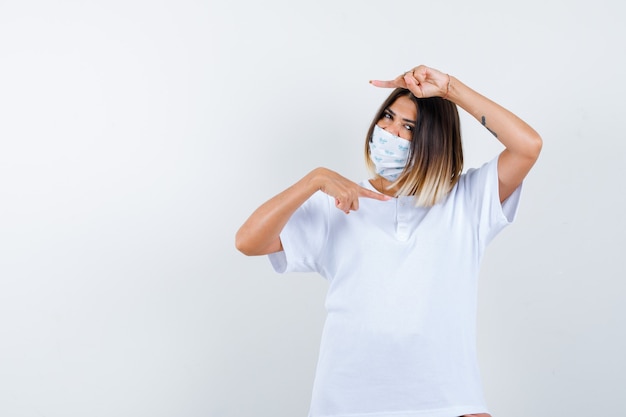 Jeune fille pointant à droite et à gauche avec l'index en t-shirt blanc et masque et à la vue de face, confiant.