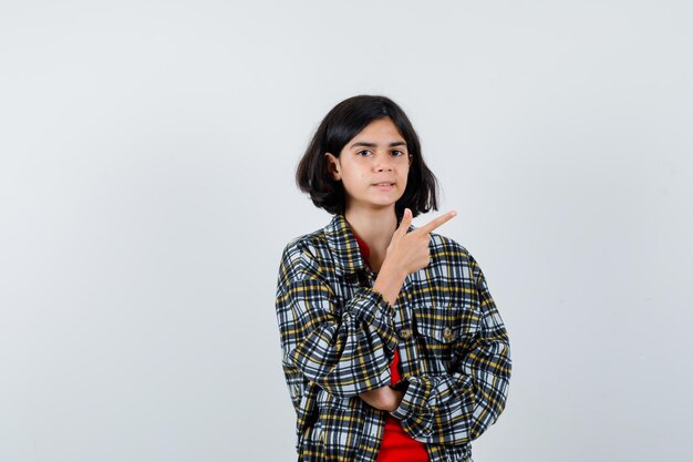 Jeune fille pointant vers la droite avec l'index en chemise à carreaux et t-shirt rouge et l'air mignon. vue de face.