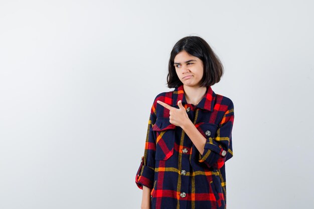 Jeune fille pointant vers la gauche en chemise à carreaux et semblant mignonne, vue de face.