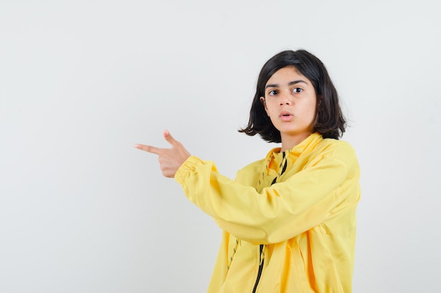 Jeune Fille Pointant Vers La Gauche Avec L'index En Bomber Jaune Et à La Surprise.