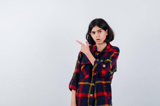 Jeune fille pointant vers la gauche avec l'index en chemise à carreaux et l'air surpris. vue de face.