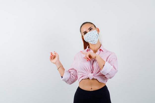 Jeune fille pointant vers la gauche avec l'index en chemisier rose, pantalon noir, masque et jolie vue de face.