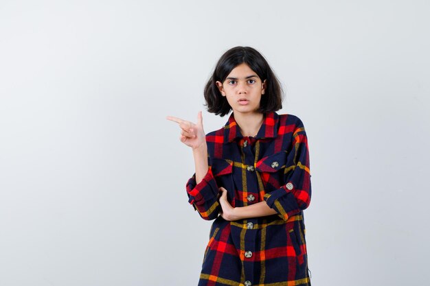 Jeune fille pointant vers la gauche tout en tenant la main sur l'avant-bras en chemise à carreaux et l'air sérieux, vue de face.