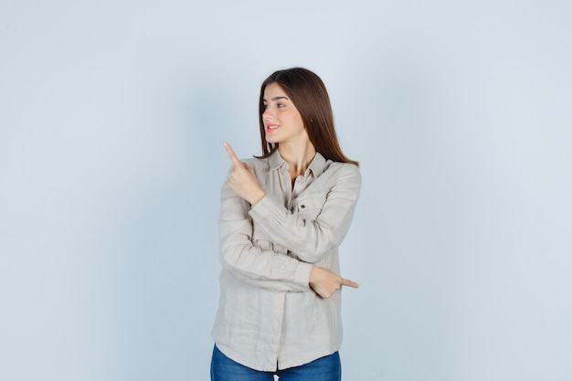 Jeune Fille Pointant Vers Le Haut Et à Droite Avec L'index En Chemise Beige, Jeans Et L'air Heureux, Vue De Face.