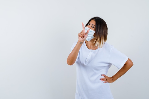 Jeune fille pointant vers le haut avec l'index, tenant la main sur la taille en t-shirt blanc et masque et à la confiance. vue de face.