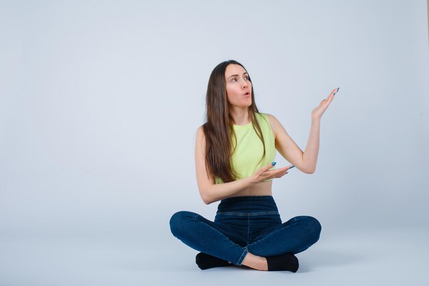 Jeune fille pointe vers la droite avec les mains en s'asseyant sur le sol sur fond blanc