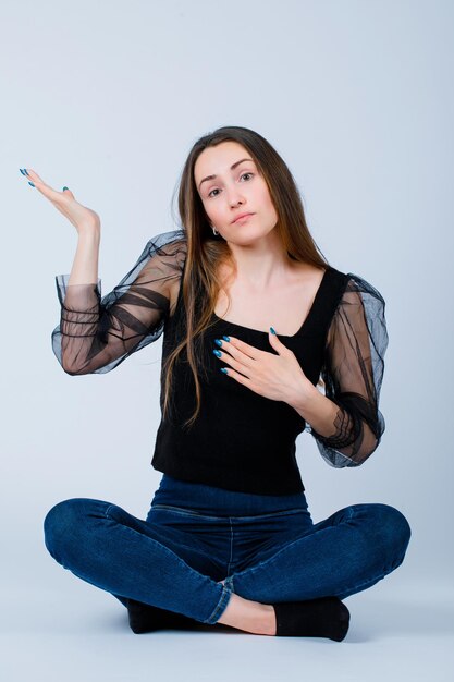 Jeune fille pointe vers la gauche avec la main en s'asseyant sur le sol sur fond blanc