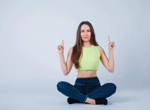 Jeune fille pointe vers le haut avec les index en s'asseyant sur le sol sur fond blanc