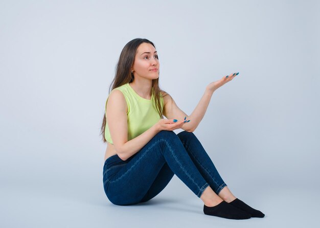 Jeune fille pointe vers le haut avec la main en s'asseyant sur le sol sur fond blanc