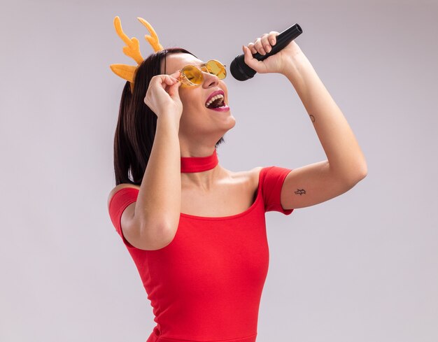 Jeune fille portant un bandeau en bois de renne et des lunettes tenant un microphone saisissant des lunettes chantant avec les yeux fermés isolés sur fond blanc