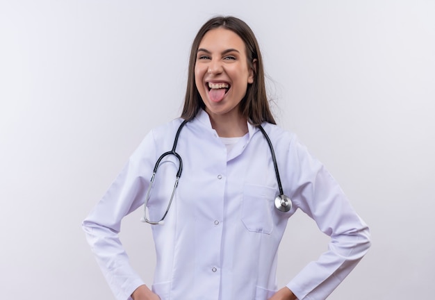 Jeune fille portant une robe médicale stéthoscope montrant la langue sur un mur blanc isolé