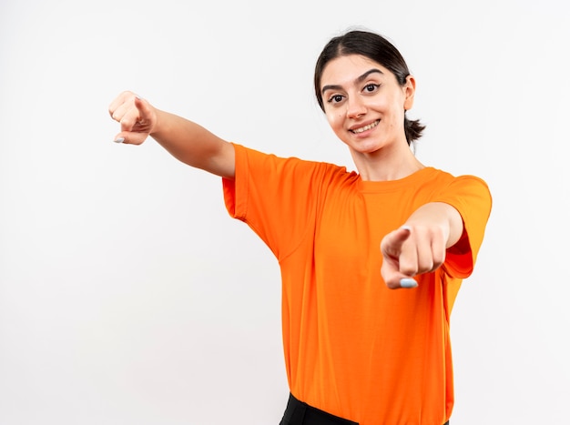 Jeune fille portant un t-shirt orange pointant avec l'index souriant joyeusement avec happy face debout sur un mur blanc