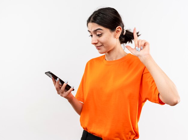 Jeune fille portant un t-shirt orange en regardant l'écran de son smartphone avec sourire sur le visage montrant l'index debout sur un mur blanc