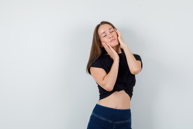 Jeune fille posant tout en touchant les joues en chemisier noir, pantalon et à la charmante.