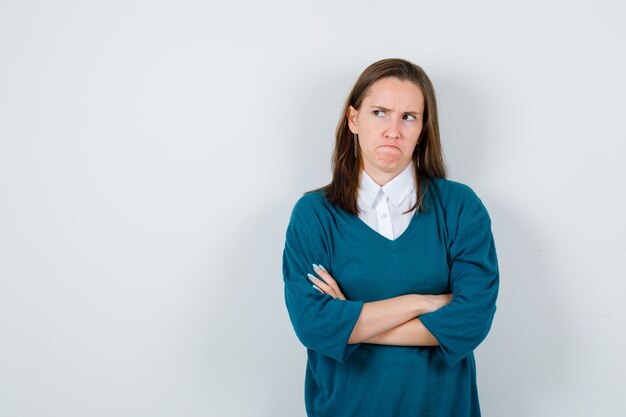 Jeune fille en pull à col en v, chemise debout avec les bras croisés, lèvres courbées et l'air lésée, vue de face.