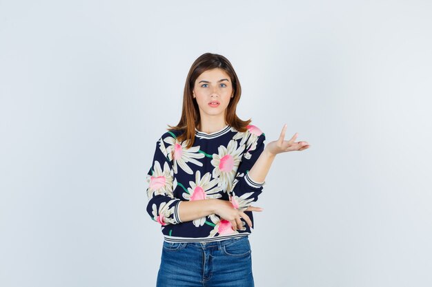 Jeune fille en pull à fleurs, jeans étirant la main de manière interrogative et l'air perplexe, vue de face.