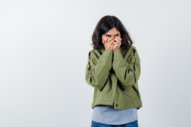 Jeune fille en pull gris, veste kaki, pantalon en jean couvrant la bouche avec les mains et l'air timide, vue de face.