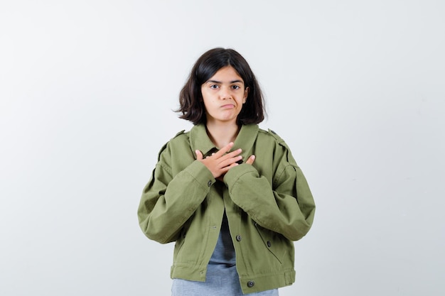 Jeune fille en pull gris, veste kaki, pantalon en jean reposant les mains sur la poitrine, grimaçant et ayant l'air sérieux, vue de face.