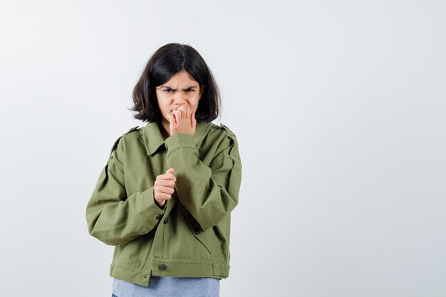 Jeune fille en pull gris, veste kaki, pantalon en jean se mordant les doigts et l'air agacé, vue de face.