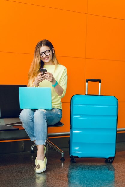 Jeune fille en pull jaune et jeans est assise sur une chaise sur fond orange. Elle a un ordinateur portable bleu sur les genoux et une valise bleue à proximité. Elle tape au téléphone.