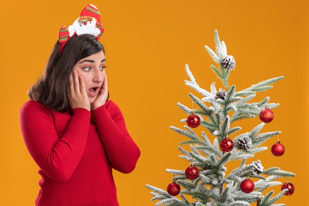 Jeune fille en pull de Noël portant un bandeau drôle à côté d'un arbre de Noël sur fond orange