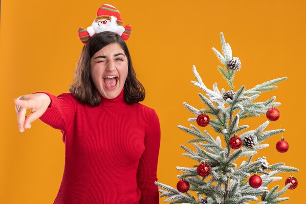 Jeune fille en pull de Noël portant bandeau drôle criant à côté d'un arbre de Noël sur fond orange