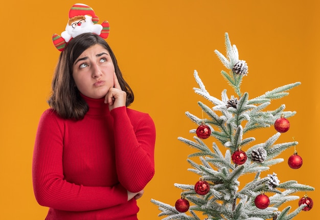 Jeune fille en pull de Noël portant un bandeau drôle à la perplexité à côté d'un arbre de Noël sur fond orange