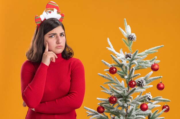 Jeune fille en pull de Noël portant un bandeau drôle regardant la caméra à côté d'un arbre de Noël sur fond orange