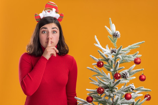 Jeune fille en pull de Noël portant un bandeau drôle regardant la caméra faisant un geste de silence à côté d'un arbre de Noël sur fond orange