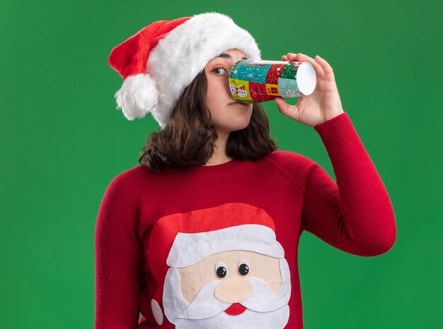 Jeune fille en pull de Noël portant bonnet de Noel de boire de la tasse de papier coloré debout sur le mur vert