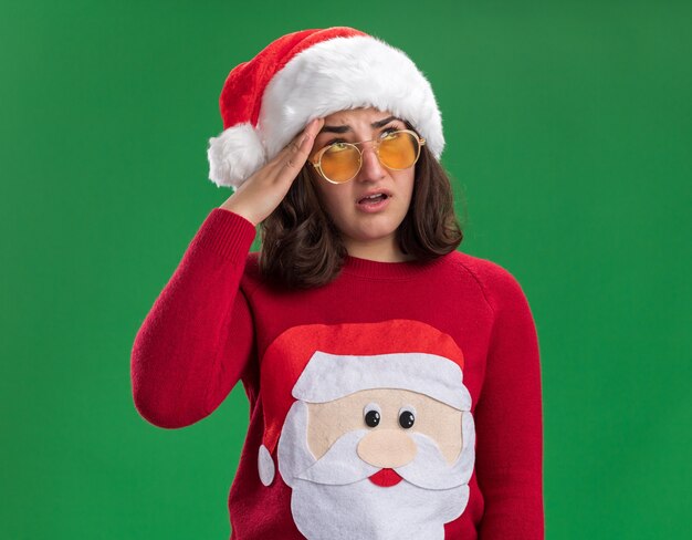 Jeune fille en pull de Noël portant bonnet de Noel et lunettes roulant les yeux fatigués et ennuyés debout sur le mur vert