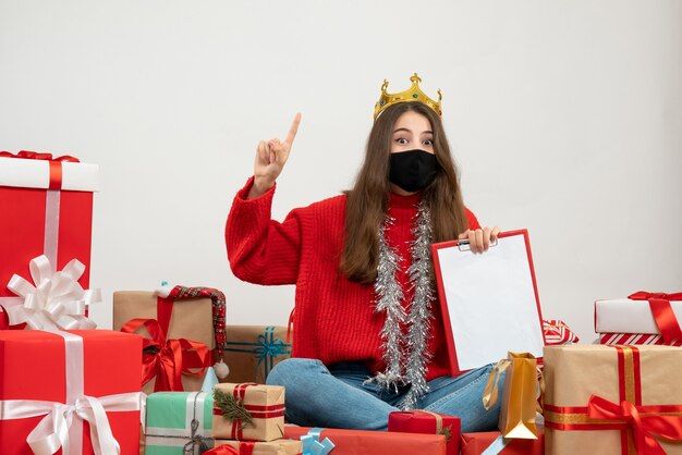 Jeune fille avec pull rouge assis autour de cadeaux avec masque noir sur blanc