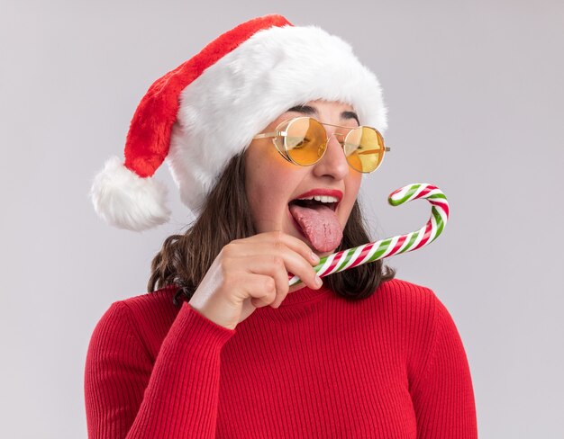 Jeune fille en pull rouge et bonnet de noel portant des lunettes tenant la canne à sucre en essayant de le goûter heureux et joyeux debout sur fond blanc