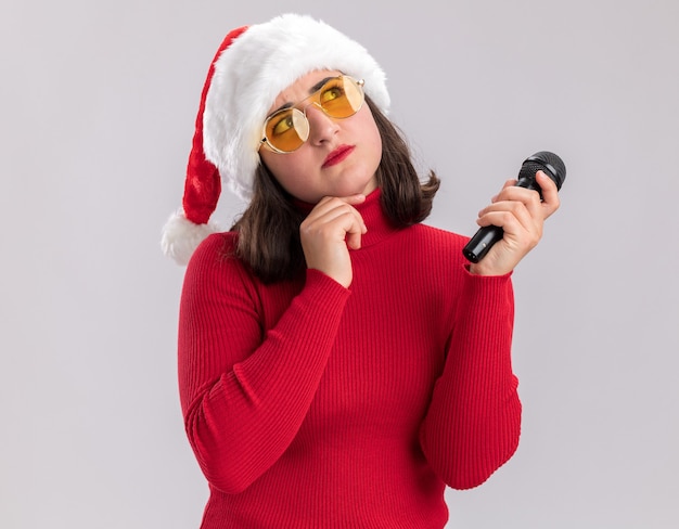 Jeune fille en pull rouge et bonnet de Noel portant des lunettes tenant le microphone à la perplexité debout sur fond blanc