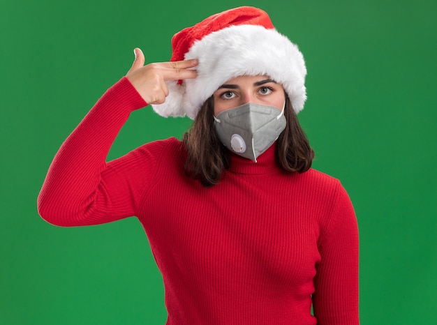 Jeune fille en pull rouge et bonnet de Noel portant un masque de protection du visage faisant le geste du pistolet avec les doigts sur sa tempe en regardant la caméra fatiguée et s'ennuie debout sur fond vert