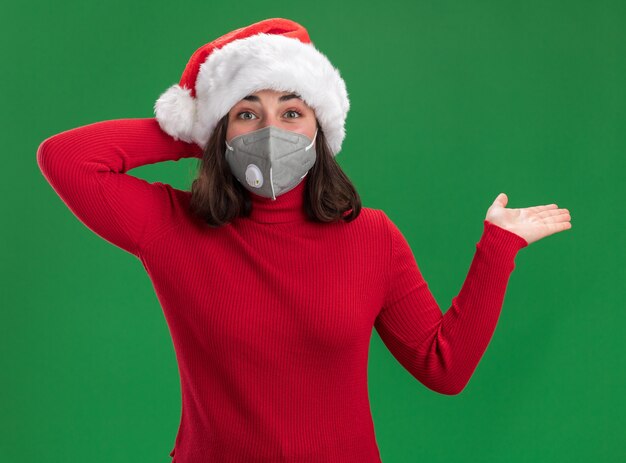 Jeune fille en pull rouge et bonnet de Noel portant un masque de protection du visage regardant la caméra heureux et surpris