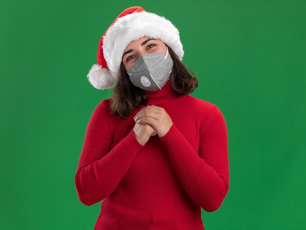 Jeune fille en pull rouge et bonnet de Noel portant un masque de protection du visage avec un visage heureux tenant les mains ensemble en attente de surprise debout sur un mur vert