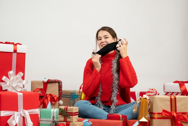 Jeune fille avec pull rouge décollant son masque assis autour de cadeaux sur blanc