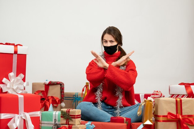 Jeune fille avec pull rouge et masque noir croisant les mains assis autour de cadeaux sur blanc