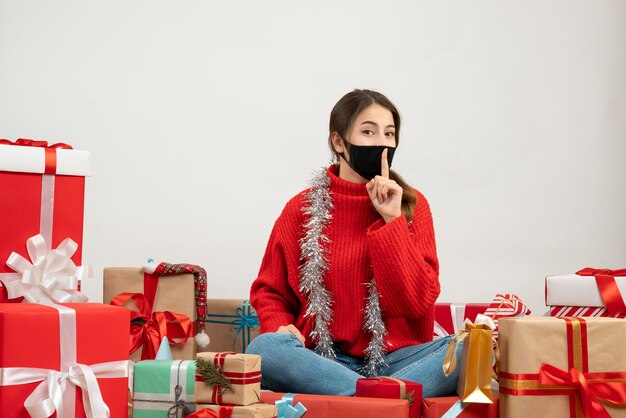 Jeune fille avec pull rouge et masque noir faisant signe chut assis autour de cadeaux sur blanc