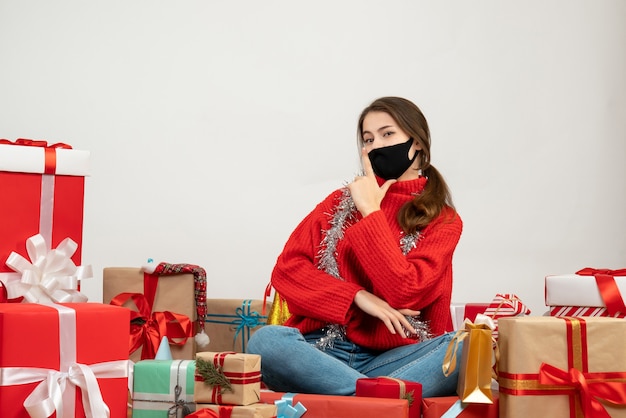 Jeune fille avec pull rouge et masque noir faisant signe de pistolet doigt assis autour de cadeaux sur blanc