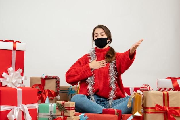 Jeune fille avec pull rouge et masque noir ouverture main fermant les yeux assis autour de cadeaux sur blanc