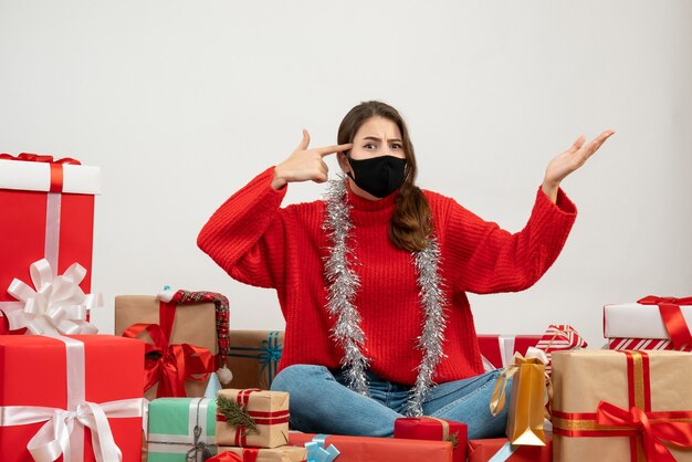 Jeune fille avec pull rouge mettant le pistolet à doigt à son temple assis autour de cadeaux avec masque noir sur blanc