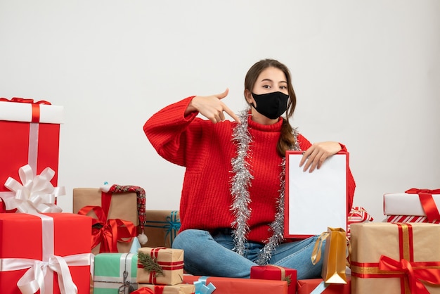 Jeune fille avec pull rouge montrant des documents assis autour de cadeaux avec masque noir sur blanc