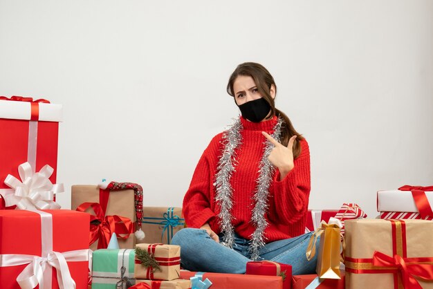 Jeune fille avec pull rouge pointant sur elle-même assis autour de cadeaux sur blanc