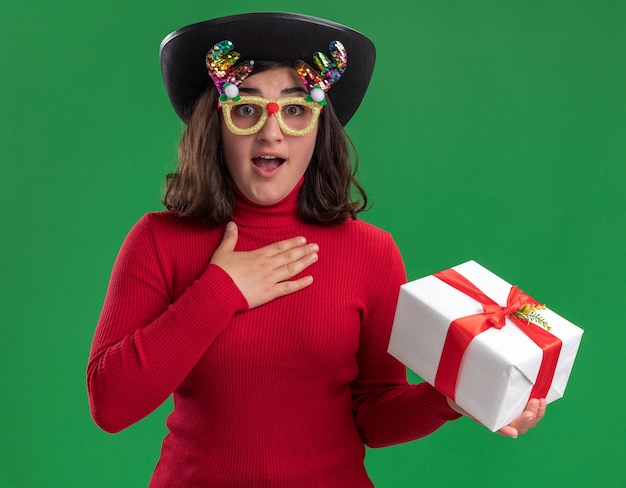 Photo gratuite jeune fille en pull rouge portant des lunettes drôles et chapeau noir tenant un cadeau regardant la caméra surpris et heureux debout sur fond vert