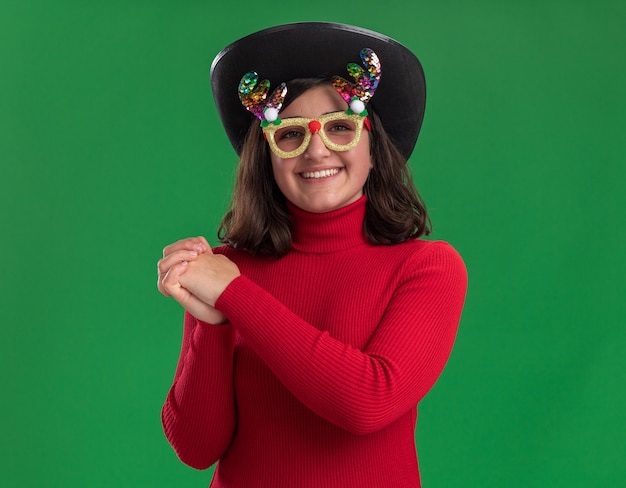 Jeune fille en pull rouge portant des lunettes drôles et chapeau noir tenant par la main ensemble heureux et positif souriant joyeusement debout sur le mur vert
