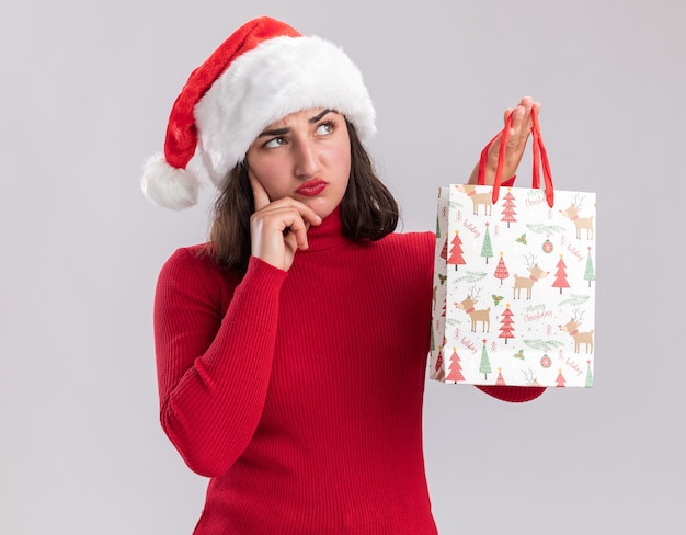 Jeune fille en pull rouge et santa hat holding sac en papier coloré avec des cadeaux de Noël à côté avec une expression sceptique pensant debout sur fond blanc